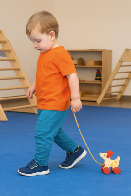 Toddler pulling a wooden Montessori dog toy across a classroom floor.